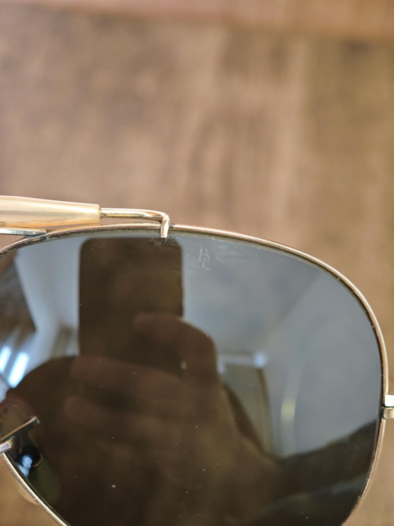 May include: Close-up of aviator sunglasses with gold-colored frames and dark lenses. The lenses reflect the surrounding environment. The top bar and temples are visible, with a light-colored temple tip.