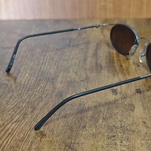 May include: Round sunglasses with dark brown lenses and a black metal frame. The glasses have a classic design with thin temples and a bridge. The lenses are set in a circular frame, offering a vintage aesthetic.