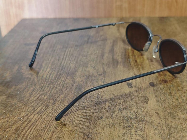 May include: Round sunglasses with dark brown lenses and a black metal frame. The glasses have a classic design with thin temples and a bridge. The lenses are set in a circular frame, offering a vintage aesthetic.