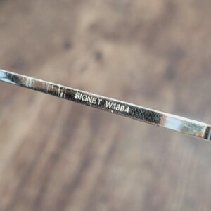 May include: Close-up of a silver eyeglass frame arm with the text "BIGNET W1384" etched on it. The frame is thin and metallic, with a reflective surface. The background is a blurred wooden surface.