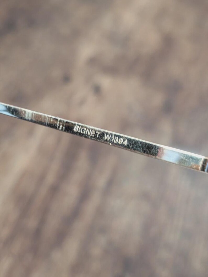 May include: Close-up of a silver eyeglass frame arm with the text "BIGNET W1384" etched on it. The frame is thin and metallic, with a reflective surface. The background is a blurred wooden surface.