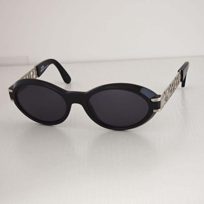 Puede incluir: Gafas de sol ovaladas con montura negra y lentes oscuras. Las patillas est&aacute;n hechas de metal plateado con un patr&oacute;n geom&eacute;trico griego repetitivo. La patilla interior muestra el texto Gianni Versace Made in Italy.