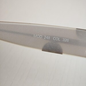 May include: A translucent, matte-finish temple arm of a pair of eyeglasses. The inner surface features the printed text MOD. 248 COL. 326 in a light, neutral font. The material has a soft, frosted appearance with a smooth, elongated shape.