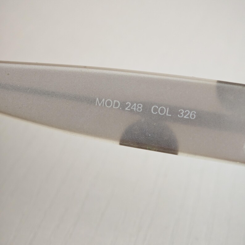 May include: A translucent, matte-finish temple arm of a pair of eyeglasses. The inner surface features the printed text MOD. 248 COL. 326 in a light, neutral font. The material has a soft, frosted appearance with a smooth, elongated shape.