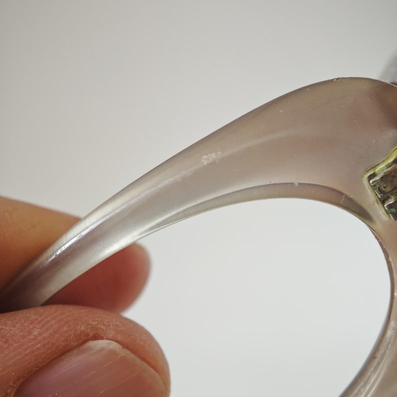 May include: A translucent, light taupe-colored curved object with a smooth, polished surface. The piece features a metallic hinge mechanism on the right side, showing a gold-toned finish with visible small screws and a jointed construction.