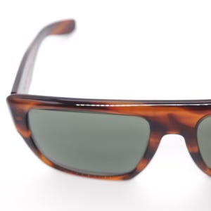 May include: Close-up of sunglasses with a tortoiseshell pattern frame and green lenses. The sunglasses have a rectangular shape and a slightly curved design. The frame is a mix of brown and amber colors.