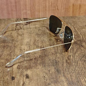 Puede incluir: Gafas de sol de aviador con montura dorada y lentes oscuras. Las gafas tienen un dise&ntilde;o cl&aacute;sico con doble puente y patillas de pl&aacute;stico transparente. Las gafas est&aacute;n sobre una superficie de madera.