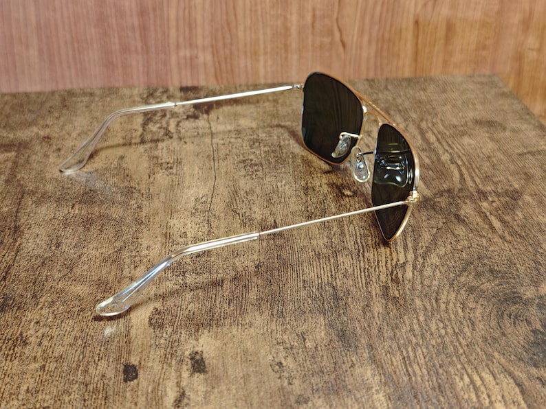 Puede incluir: Gafas de sol de aviador con montura dorada y lentes oscuras. Las gafas tienen un dise&ntilde;o cl&aacute;sico con doble puente y patillas de pl&aacute;stico transparente. Las gafas est&aacute;n sobre una superficie de madera.