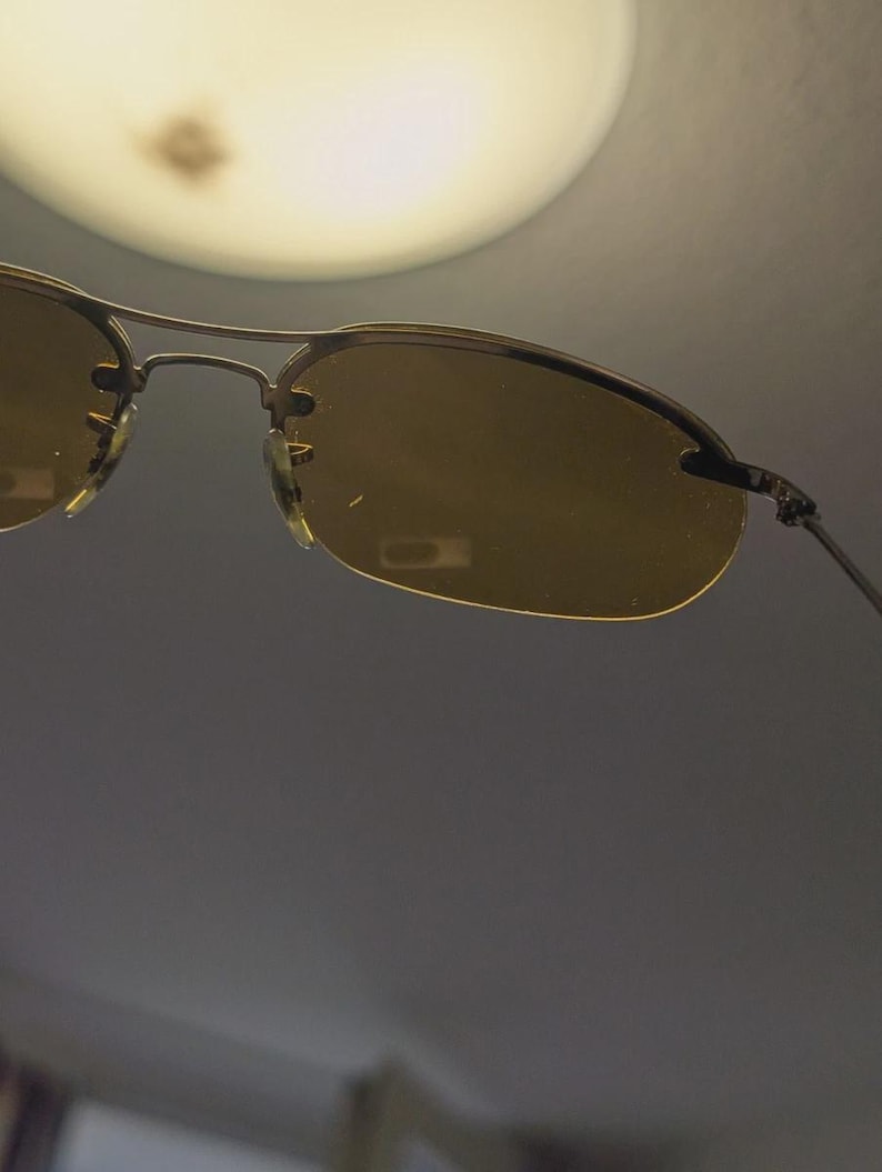 May include: Rimless sunglasses with a gold-colored metal frame and amber-tinted lenses. The sunglasses have a classic design with a double bridge and thin temples. The lenses are oval-shaped and provide a warm, golden hue.
