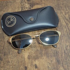 May include: Gold-framed sunglasses with dark lenses and a black leather case. The sunglasses have a classic oval shape and a decorative design on the temples. The case features the Ray-Ban logo.