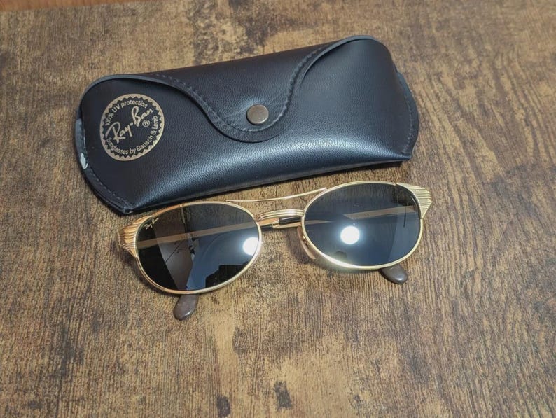 May include: Gold-framed sunglasses with dark lenses and a black leather case. The sunglasses have a classic oval shape and a decorative design on the temples. The case features the Ray-Ban logo.