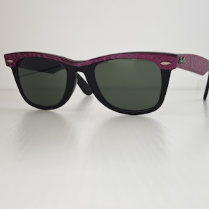 Puede incluir: Gafas de sol con montura negra, barra superior y patillas texturizadas en tonos morados. Las lentes son de color verde oscuro y la montura incluye detalles met&aacute;licos plateados en las esquinas exteriores cerca de las bisagras.