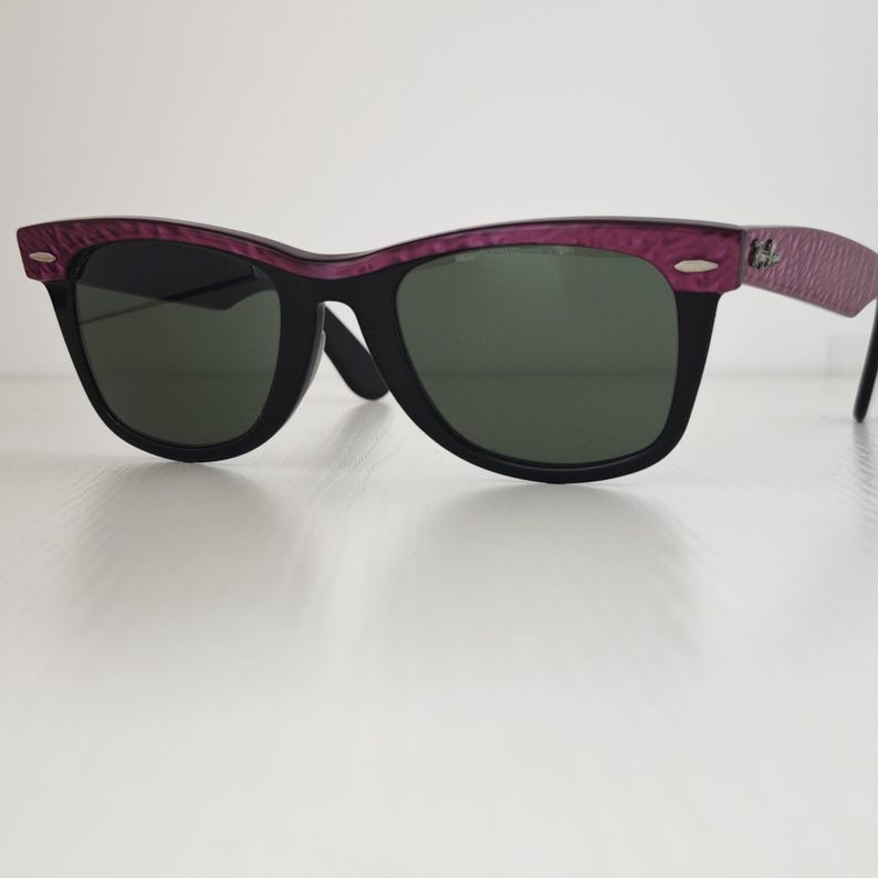 Puede incluir: Gafas de sol con montura negra, barra superior y patillas texturizadas en tonos morados. Las lentes son de color verde oscuro y la montura incluye detalles met&aacute;licos plateados en las esquinas exteriores cerca de las bisagras.