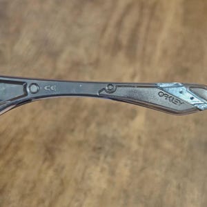 May include: Close-up of a silver-toned Oakley sunglass arm. The arm features the brand name "Oakley" and the letters "CE". The arm has a geometric design near the hinge.