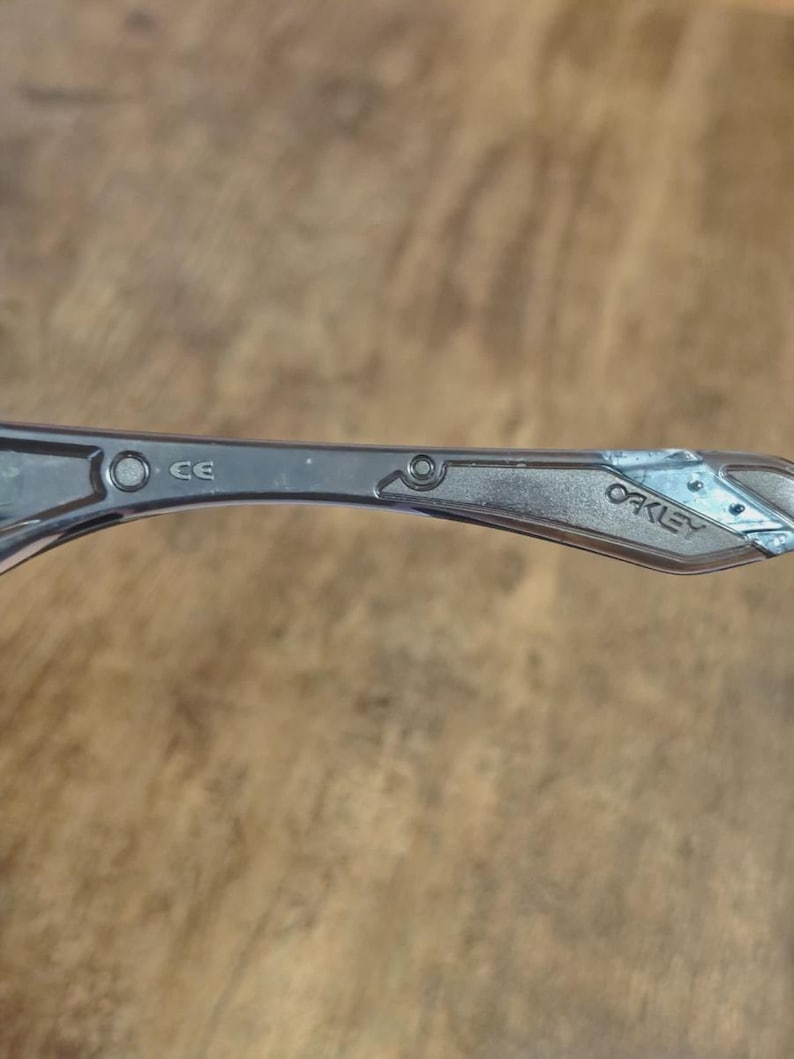 May include: Close-up of a silver-toned Oakley sunglass arm. The arm features the brand name "Oakley" and the letters "CE". The arm has a geometric design near the hinge.