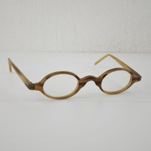 May include: A pair of spectacles featuring oval-shaped frames with a light brown and tan marbled pattern. The glasses have a high bridge and thin, matching temples that extend outwards, showcasing a classic design with a smooth, polished finish.