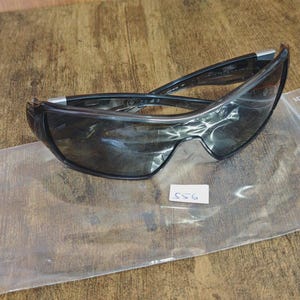 May include: Black sunglasses with a curved design, resting on a clear plastic bag. The lenses are dark, and the frames are black with silver accents. A small white tag with the number "556" is visible.