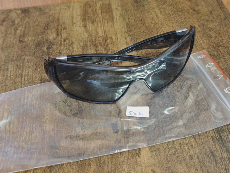 May include: Black sunglasses with a curved design, resting on a clear plastic bag. The lenses are dark, and the frames are black with silver accents. A small white tag with the number "556" is visible.