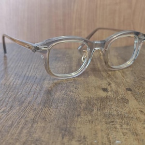 Puede incluir: Gafas con montura transparente de forma rectangular. Las monturas son de color gris claro translúcido y las lentes son transparentes. Las patillas son de color marrón claro. Las gafas están sobre una superficie de madera.