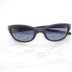 May include: Black sunglasses with a curved frame and dark lenses. The brand name "Oakley" is visible on the frame. The sunglasses are set against a white background.