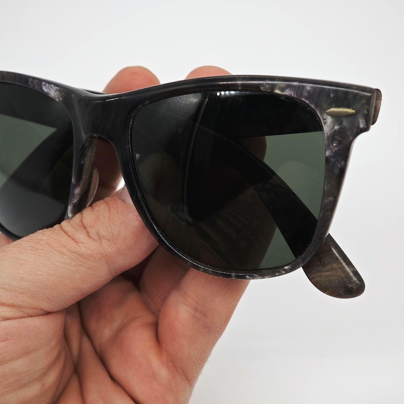 Puede incluir: Primer plano de gafas de sol con montura veteada en negro y gris y lentes verde oscuro. Las gafas de sol tienen una forma rectangular cl&aacute;sica y est&aacute;n sujetas por una mano sobre un fondo blanco. Las patillas son de un tono gris m&aacute;s oscuro.