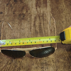 May include: Gold-framed sunglasses with dark lenses, measured at approximately 5 inches in length. The sunglasses are positioned on a wooden surface, with a measuring tape alongside them.