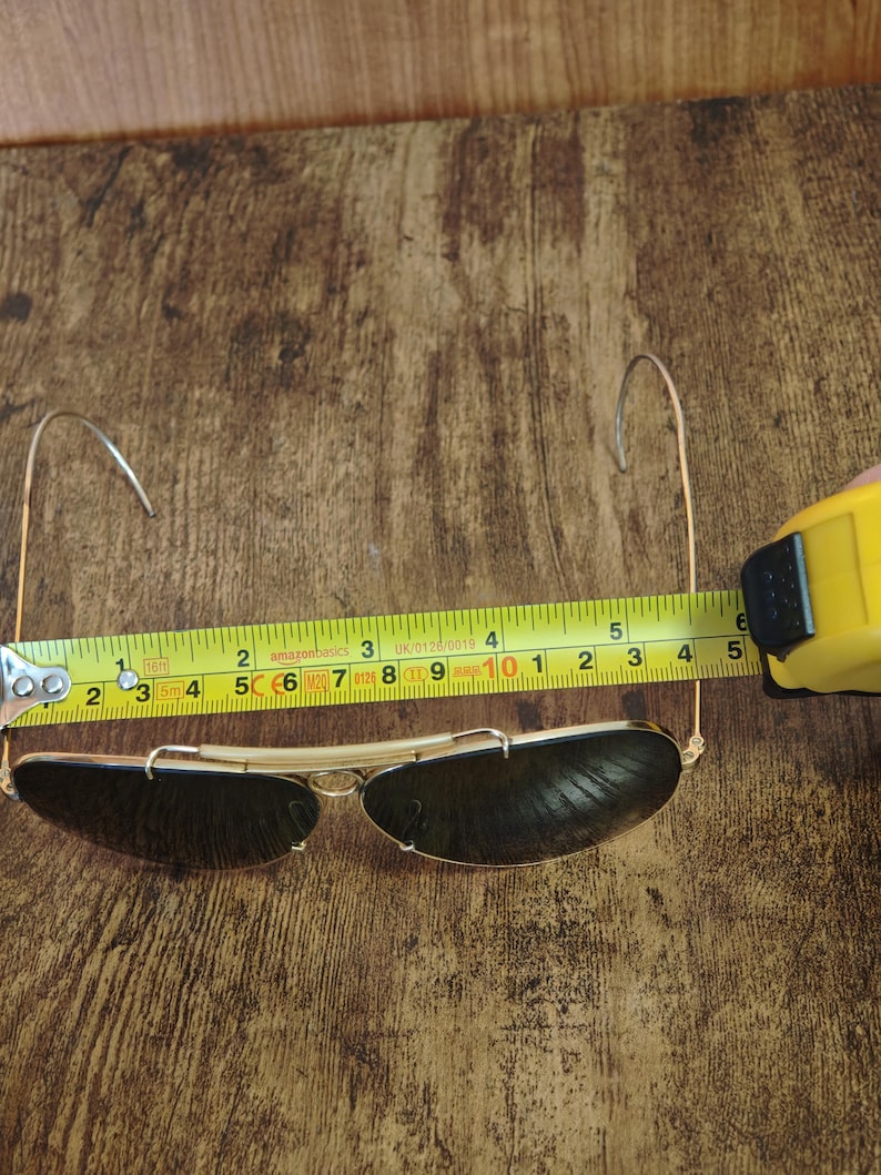 May include: Gold-framed sunglasses with dark lenses, measured at approximately 5 inches in length. The sunglasses are positioned on a wooden surface, with a measuring tape alongside them.