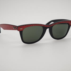 May include: Black sunglasses with a red patterned frame and dark green lenses. The sunglasses have a classic rectangular shape. The temples are red with a textured pattern and a small emblem.