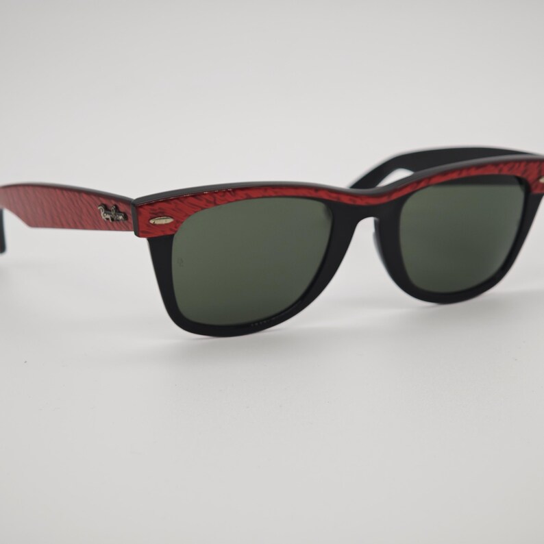 May include: Black sunglasses with a red patterned frame and dark green lenses. The sunglasses have a classic rectangular shape. The temples are red with a textured pattern and a small emblem.