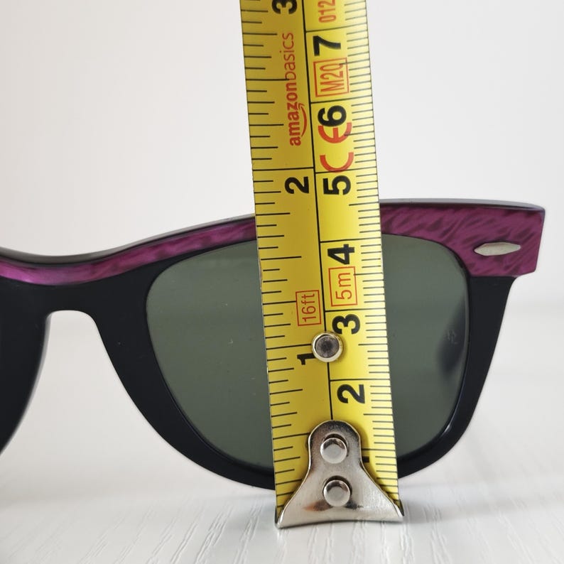 Puede incluir: Gafas de sol negras con una parte superior texturizada en color morado y lentes verde oscuro. Una cinta m&eacute;trica amarilla est&aacute; colocada verticalmente sobre el centro de la montura, mostrando una altura de lente de unos 4,5 cm.