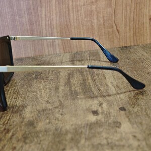 Peut inclure: Lunettes de soleil noires avec des branches dor&eacute;es et des verres fonc&eacute;s. Les lunettes ont une monture rectangulaire. Les branches sont fines et se prolongent par des embouts noirs. Fond en bois.