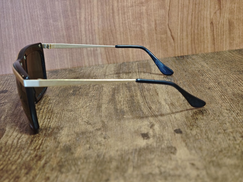 Peut inclure: Lunettes de soleil noires avec des branches dor&eacute;es et des verres fonc&eacute;s. Les lunettes ont une monture rectangulaire. Les branches sont fines et se prolongent par des embouts noirs. Fond en bois.