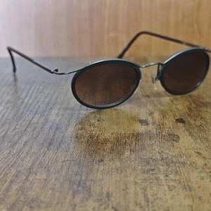 May include: Oval-shaped sunglasses with dark brown lenses and a thin, dark metal frame. The sunglasses are resting on a wooden surface, showcasing their sleek design and vintage aesthetic.