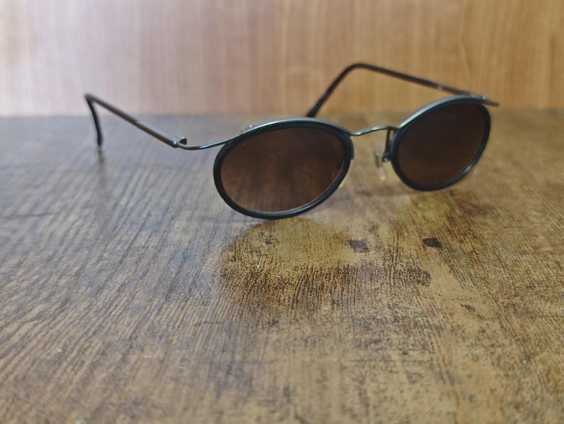 May include: Oval-shaped sunglasses with dark brown lenses and a thin, dark metal frame. The sunglasses are resting on a wooden surface, showcasing their sleek design and vintage aesthetic.