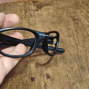 Op de afbeelding: Zwarte bril met een sportief design. De monturen hebben een matte afwerking en een getextureerde grip op de pootjes. De glazen zijn helder en de merknaam is zichtbaar op het montuur.