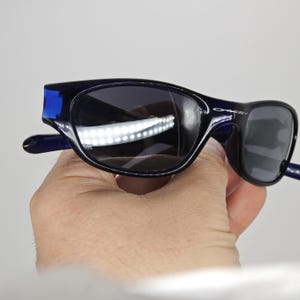 May include: Dark blue Oakley sunglasses with black lenses. The sunglasses have a curved frame and the Oakley logo is visible on the right arm. The sunglasses are held in a hand.