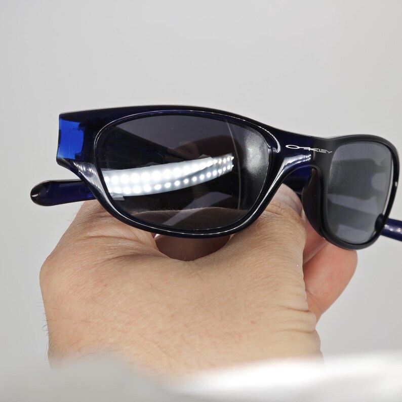 May include: Dark blue Oakley sunglasses with black lenses. The sunglasses have a curved frame and the Oakley logo is visible on the right arm. The sunglasses are held in a hand.