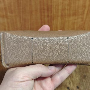 May include: A tan rectangular eyeglasses case with a textured surface. The case has three vertical black lines on the top. The case is held in a hand.
