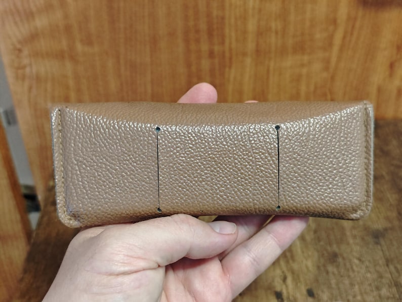 May include: A tan rectangular eyeglasses case with a textured surface. The case has three vertical black lines on the top. The case is held in a hand.