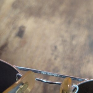 May include: Close-up of a pair of sunglasses with a silver frame and dark lenses. The text "B&L RAY-BAN" is visible on the frame. The nose pads are a light brown color.