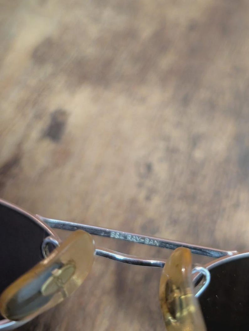 May include: Close-up of a pair of sunglasses with a silver frame and dark lenses. The text "B&L RAY-BAN" is visible on the frame. The nose pads are a light brown color.