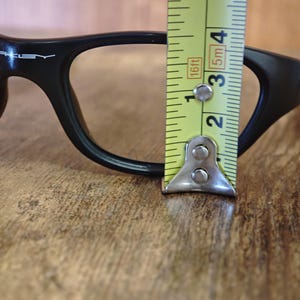 Op de afbeelding: Zwarte bril met een meetlint dat een breedte van ongeveer 5 cm aangeeft. De bril heeft een strak, modern design met de merknaam "Oakley" zichtbaar op het montuur. De achtergrond is een houten oppervlak.