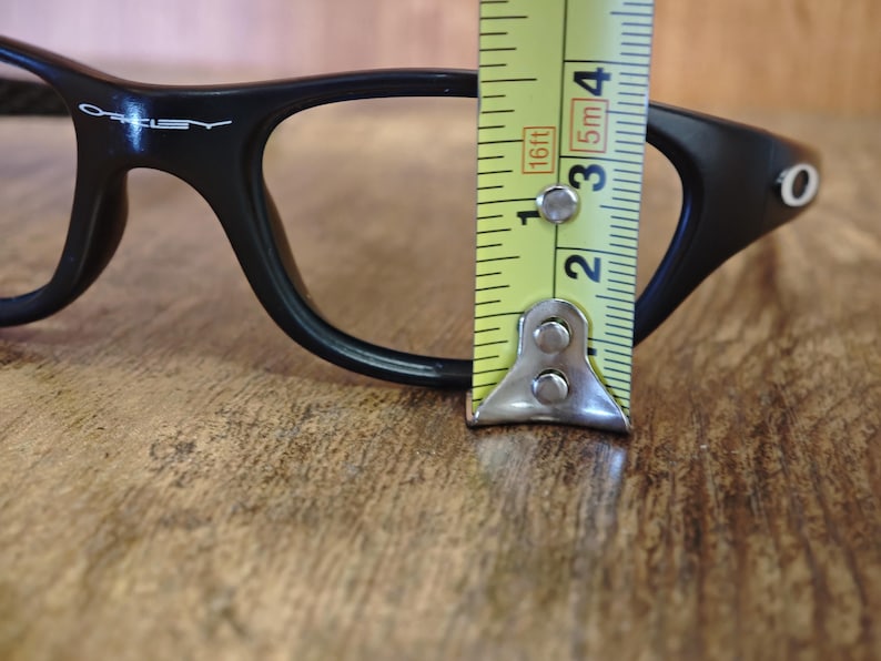 Op de afbeelding: Zwarte bril met een meetlint dat een breedte van ongeveer 5 cm aangeeft. De bril heeft een strak, modern design met de merknaam "Oakley" zichtbaar op het montuur. De achtergrond is een houten oppervlak.