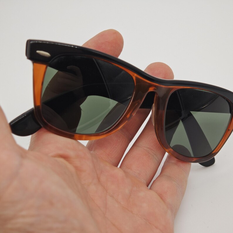 May include: Sunglasses with a black frame and orange-brown accents. The lenses are dark green, and the sunglasses are held in a hand. The sunglasses have a classic, retro design.