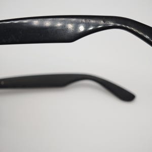 May include: Close-up of black sunglasses arms. The arms curve downward and have a smooth, matte finish. The top arm shows signs of wear. The sunglasses are set against a white background.