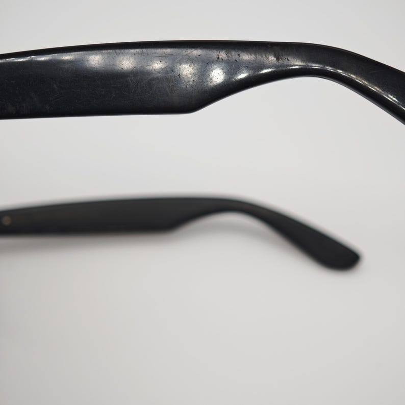 May include: Close-up of black sunglasses arms. The arms curve downward and have a smooth, matte finish. The top arm shows signs of wear. The sunglasses are set against a white background.