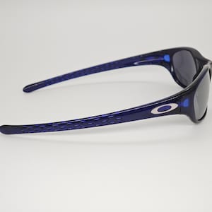 May include: Navy blue sunglasses with a contemporary design. The frames are a dark blue with a textured pattern on the arms. The lenses are a dark grey, contrasting with the blue frames. The Oakley logo is visible on the side of the sunglasses.