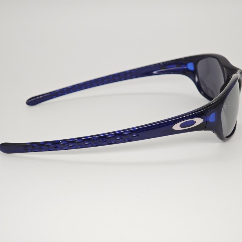 May include: Navy blue sunglasses with a contemporary design. The frames are a dark blue with a textured pattern on the arms. The lenses are a dark grey, contrasting with the blue frames. The Oakley logo is visible on the side of the sunglasses.