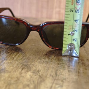 May include: Brown tortoiseshell sunglasses with dark lenses. The sunglasses are approximately 2 inches (5 cm) tall. The Ray-Ban logo is visible on the left lens. The sunglasses are on a wooden surface.