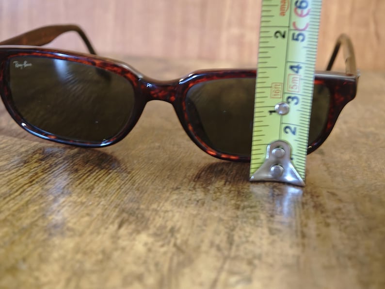 May include: Brown tortoiseshell sunglasses with dark lenses. The sunglasses are approximately 2 inches (5 cm) tall. The Ray-Ban logo is visible on the left lens. The sunglasses are on a wooden surface.